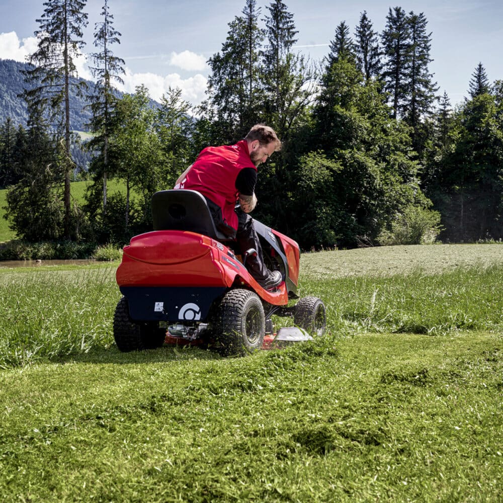 Mand kører plænetraktor på en græsmark med skov baggrund.