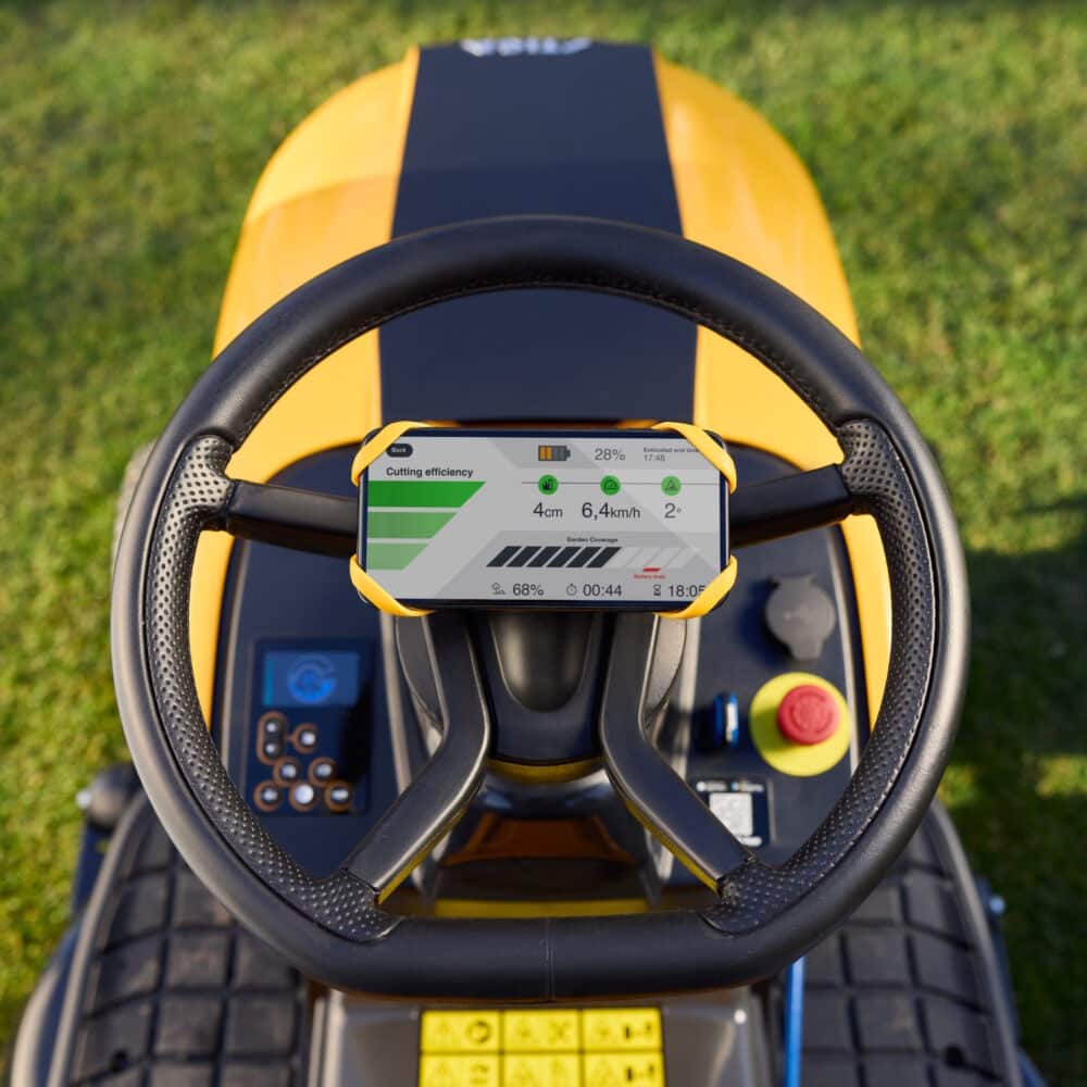 En gul græsslåmaskine set fra oven, hvor der er monteret en smartphone på rattet. Telefonen viser oplysninger om klippeeffektivitet, inklusive højde på 4 cm, hastighed på 6,4 km/t, og batteriniveau. Græsplænen ses i baggrunden.