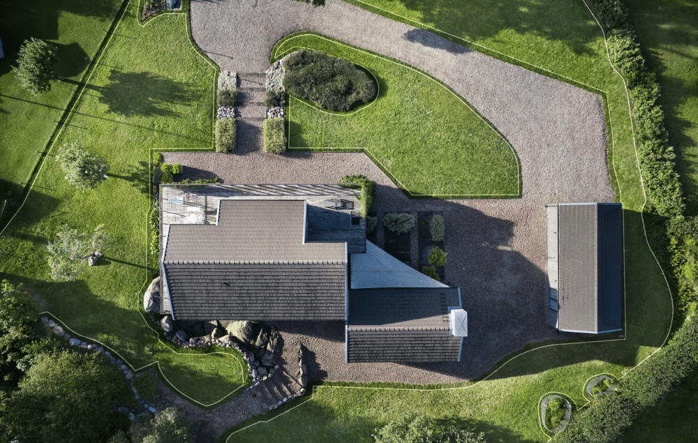 Luftfoto af et hus omgivet af grønne plæner og en grusbelagt gårdsplads.