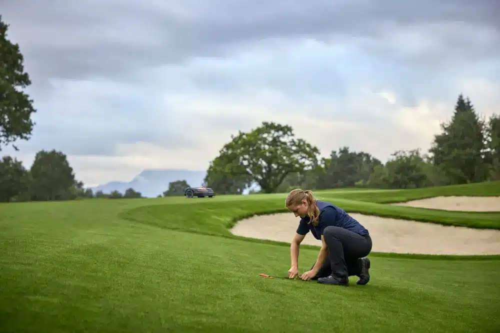 En person knæler på en frodig grøn golfbane og arbejder med jorden. I baggrunden er der et sandbunker og et robotgræsslåmaskine, omgivet af træer og en overskyet himmel.