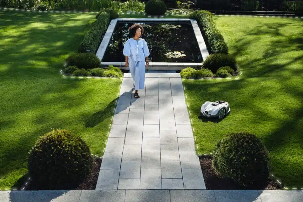 Kvinde går forbi en robotplæneklipper i en have.