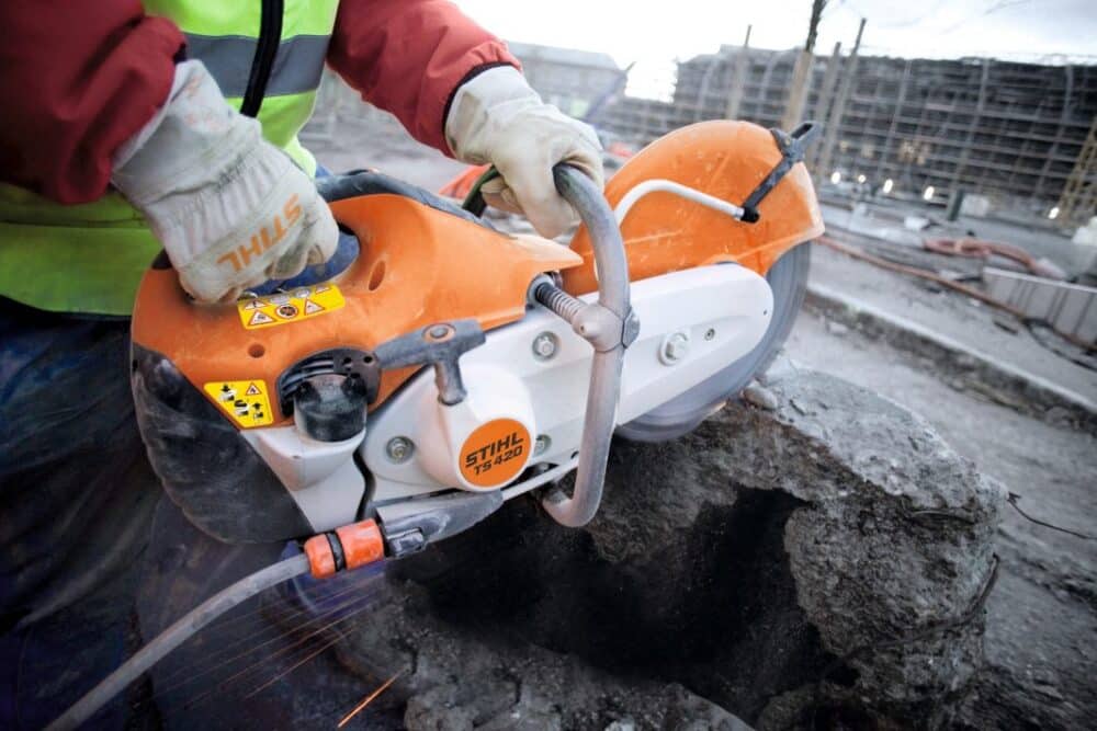 Arbejdsperson iført sikkerhedsvest og handsker bruger en orange STIHL TS 420 skæresav til at skære i beton på en byggeplads.
